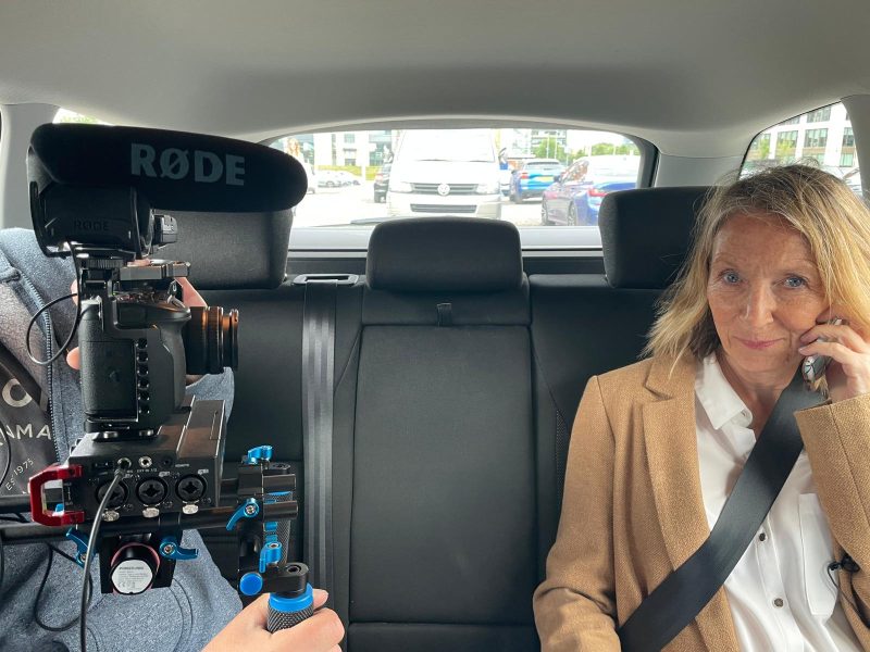 Behind-the-scenes shot of a video production crew filming a woman sitting in the backseat of a car while talking on the phone, with professional camera and microphone equipment visible.